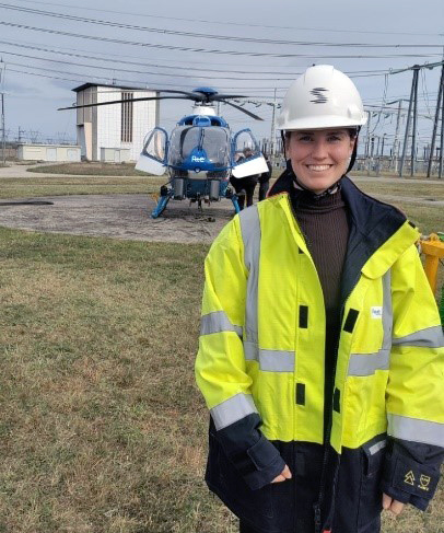 Greta Jonsson i skyddshjälm och varselväst, framför en helikopter och en kraftstation med kraftledningar.