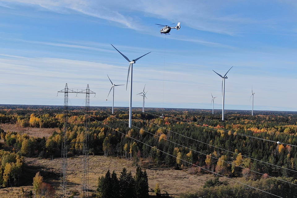Bilden visar en helikopter som flyger i luften i området över Hallstavik. Trädtopparna är gula och bruna och gröna, det är höst. Kraftledningen syns tydligt över trädtopparna genom terrängen. Vid kraftledningen är flyghindermarkörer uppsatta.