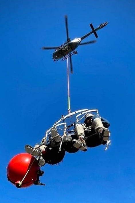 Blå himmel är bakgrund, man ser en helikopter som hoovrar i luften, ifrån helikopterns medar hänger den särskilda korgen som används för att fästa flyghinderklot vid kraftledningen.