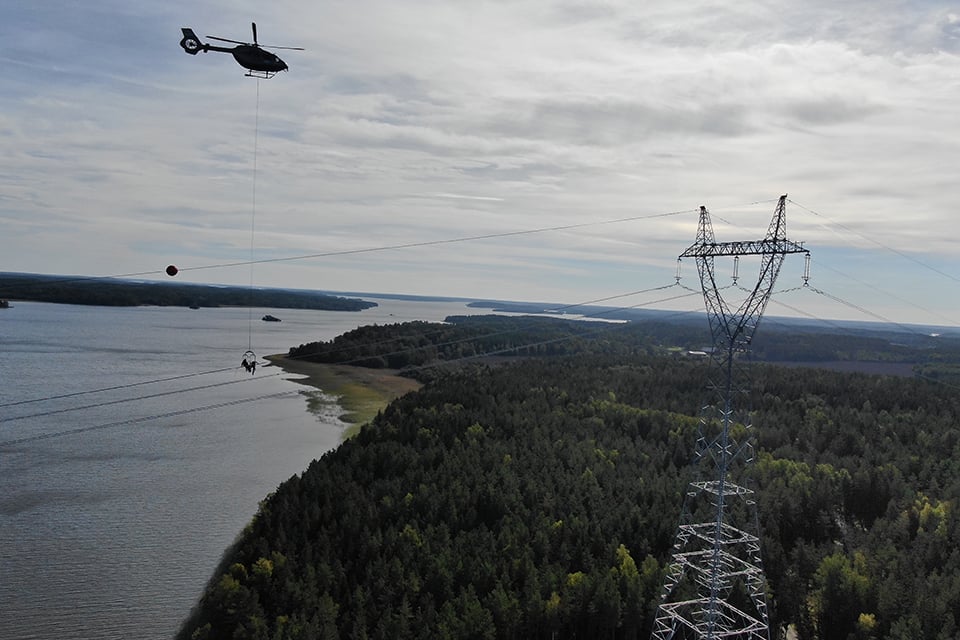 Bilden visar några av Svenska kraftnäts högsta stolpar, de så kallade Mälartornen som med en tydlig majestätisk stolprofil reser sig ur terrängen. Vatten och natur syns på marken, längre ned. På kraftledningen hänger flyghindermarkörer.