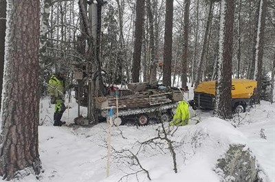 En borrvagn i skog och snö. En gulklädd person står bredvid borrvagnen.