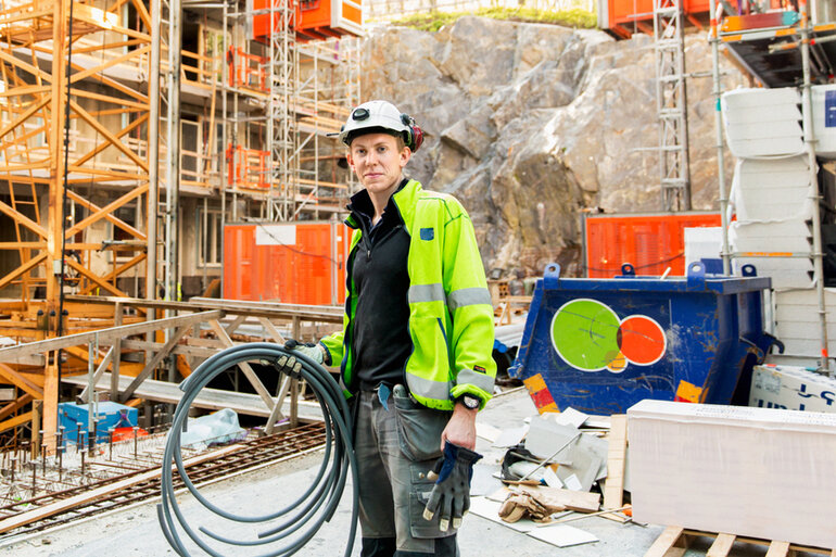 En manlig elektriker står på ett bygge, han har gul varselväst och en vit hjälm och håller plaströr i ena handen.