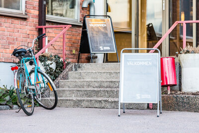 Ingång till byggnad där det finns en skylt där det står välkommen. Det finns också cyklar utanför.