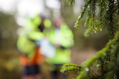 Oskarp bild på två personer som står och pratar i skogen klädda i varselkläder