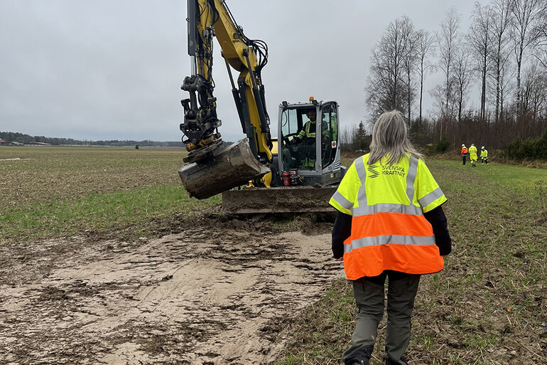 På en öppen markyta håller en grävmaskin på att gräva bort det översta lagret jord. I förgrunden syns en person i Svenska kraftnät-jacka.